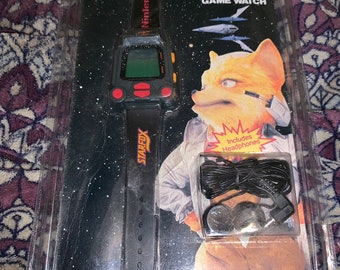 May include: A black Nintendo Star Fox Game Watch with a black wristband and a yellow button. The watch is packaged in a clear plastic case with a black and white image of Star Fox on the back. The packaging also includes a pair of black headphones.