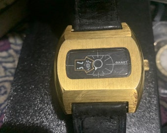 May include: A vintage gold-toned watch with a rectangular face and a black leather band. The watch face displays the brand name "GANET" and a unique time display. The watch has a classic, retro aesthetic.