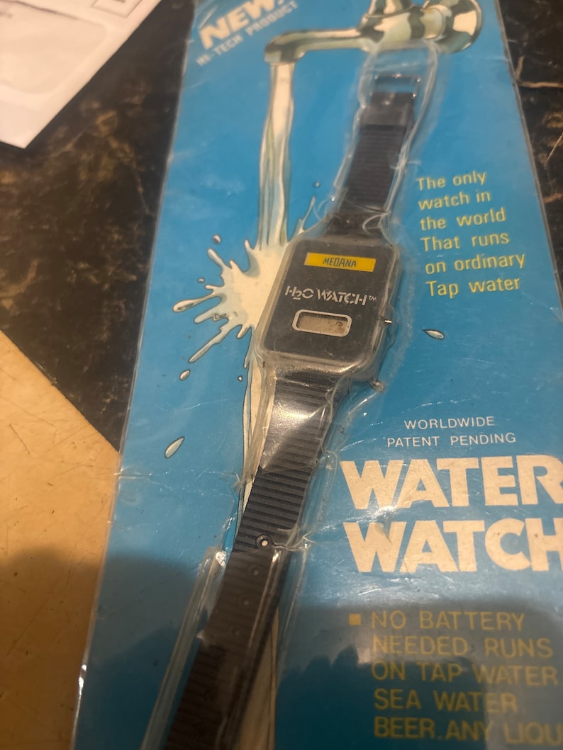 May include: A black and silver water-powered watch on a blue and white package. The package says "NEW HI-TECH PRODUCT" and "WORLDWIDE PATENT PENDING WATER WATCH". The watch has a digital display and the text "MEDANA H₂O WATCH&trade;". The package also says "The only watch in the world That runs on ordinary Tap water" and "NO BATTERY NEEDED RUNS ON TAP WATER SEA WATER BEER.ANY LIQU".