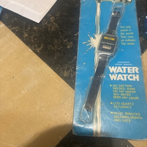May include: A blue and white packaging card for a water-powered watch. The watch is black with a digital display and the text "H₂O WATCH" is printed on the face. The packaging card features a cartoon illustration of a faucet with water flowing from it. The text "NEW! HI-TECH PRODUCT" is printed at the top of the card. The text "WORLDWIDE PATENT PENDING WATER WATCH" is printed in large letters at the bottom of the card. The text "NO BATTERY NEEDED. RUNS ON TAP WATER SEA WATER BEER.ANY LIQUID" is printed in a smaller font below the large text. The text "LCD QUARTZ ACCURACY" is printed below the previous text. The text "HOUR. MINUTES. SECONDS.MONTH AND DATE" is printed at the bottom of the card.