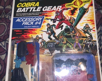 May include: A G.I. Joe Cobra Battle Gear Accessory Pack #4 box with a red and blue background. The box features a Cobra logo and the text "Cobra Battle Gear". The box contains a variety of accessories for use with Cobra figures, including weapons, backpacks, and battle stands. The box also includes an exclusive missile system.