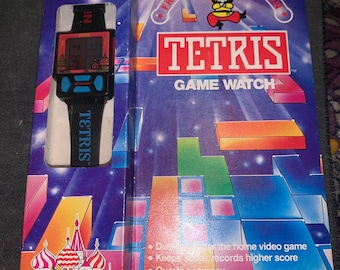 May include: A blue and red box with a picture of a Tetris game watch on the front. The box is labeled "Nintendo Tetris Game Watch by Nelsonic". The back of the box has a picture of a Russian city and a list of features. The box is labeled "Official Nintendo Licensed Product".