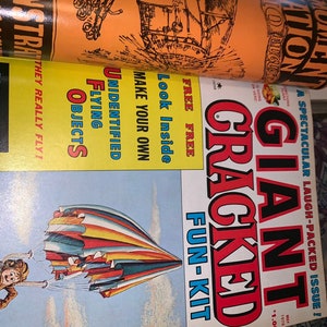 May include: A vintage magazine with the title "Giant Cracked Fun-Kit" in red and black, featuring a colorful parachute illustration. Other text includes "Unidentified Flying Objects" and "Demonstration".