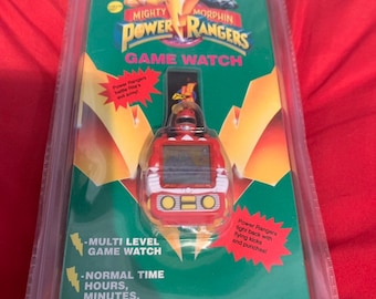 May include: A red and yellow Power Rangers Game Watch with a multi-level game watch and normal time features. The watch features a red Power Ranger character on the face. The packaging includes the text "Mighty Morphin Power Rangers Game Watch".