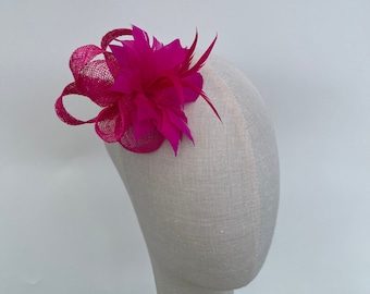 May include: A fuchsia-coloured fascinator with a floral and feather arrangement. The headpiece is mounted on a mesh base with a looped detail. The accessory is designed to be worn on the side of the head.