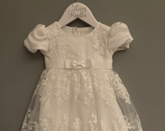 Puede incluir: Vestido blanco de bebé con detalles de encaje y un lazo de satén. El vestido está en una percha con el texto "Loopy Roodles" en él.