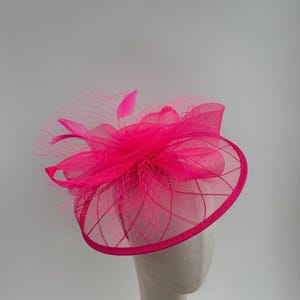 May include: A bright fuchsia fascinator with a circular base and a fine net overlay. The headpiece features a central floral design and feather accents, suitable for special events. The hat is displayed on a mannequin head.