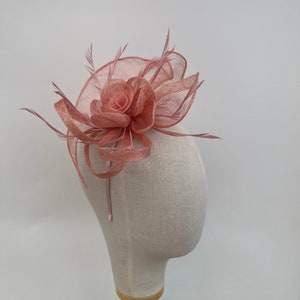 May include: A blush pink fascinator with a central rose-shaped design and feather detailing. The headpiece is made from a woven fabric, with a circular base and decorative elements. This formal headwear is suitable for special occasions such as weddings or the races.