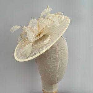 May include: A cream-colored fascinator with a circular base and a large, curled feather design. The fascinator is made of a delicate, woven material.