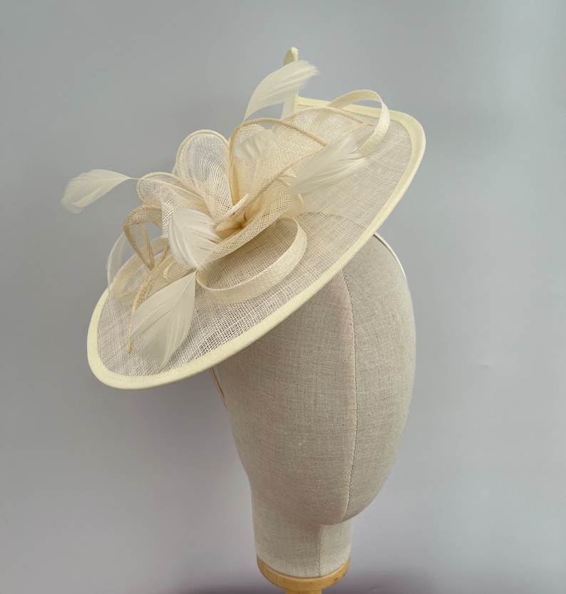 May include: A cream-colored fascinator with a circular base and a large, curled feather design. The fascinator is made of a delicate, woven material.