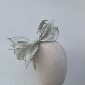 May include: A silver headpiece featuring a large bow and feather detailing. The bow is crafted from a textured, shimmering material. The headpiece is displayed on a mannequin head, highlighting its form. This accessory is suitable for special events and celebrations.