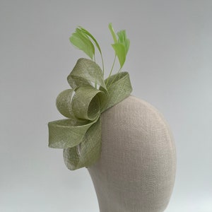 May include: A light green fascinator with a bow and feathers. The headpiece is made of woven material and features a large, looped bow on the side. Several light green feathers extend upwards from the bow.