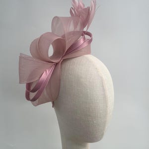 May include: A dusky pink fascinator featuring a large bow and feather detailing. The headpiece is displayed on a mannequin head. The bow is crafted from sheer material, and the feathers are in a complementary shade of pink.