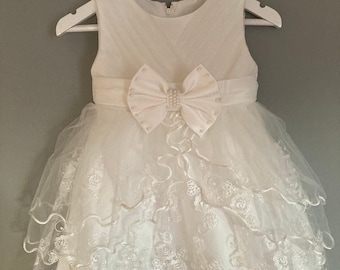 Puede incluir: Vestido blanco sin mangas con falda de tul y ribete de encaje. El vestido tiene un lazo de satén blanco con adornos de perlas y una cremallera en la espalda. El vestido está colgado en una percha blanca con el texto "Livvy Roodles" escrito en ella.