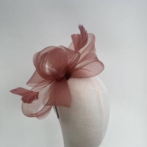 May include: A mauve headpiece featuring a large, layered mesh bow and matching feathers. The headpiece is displayed on a mannequin head, highlighting its design. This accessory is likely for special events or formal occasions.