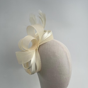 May include: A cream-coloured fascinator featuring a looped ribbon design and feather accents. The headpiece is attached to a fabric-covered base, creating an elegant look. Ideal for weddings or special occasions.