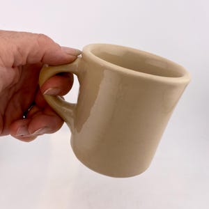 May include: A white ceramic mug with a black handle and the text "But First, Coffee" in a bold, black font. The mug is set against a blurred background, highlighting its clean design.