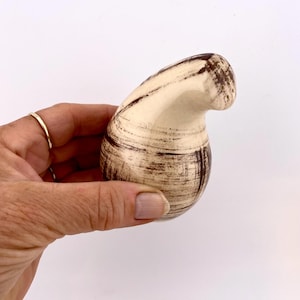 May include: A ceramic salt shaker with a cream-colored base and dark brown brushstroke design. The shaker has a curved, organic shape and a small opening at the top. The design is reminiscent of a mushroom.