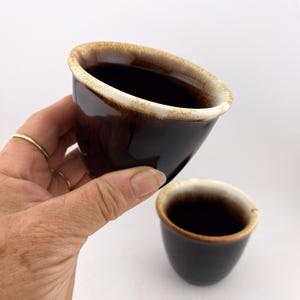 May include: Two small, dark brown ceramic cups with a glossy finish. The rims have a light brown and white glaze. One cup is held in a hand, and the other is resting on a white surface. The cups have a simple, minimalist design.