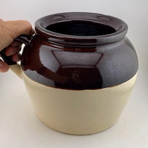 May include: A two-tone ceramic crock with a handle. The upper portion is a glossy dark brown, while the lower section is a creamy beige. The crock has a wide opening and a rounded body, suitable for kitchen or home decor.