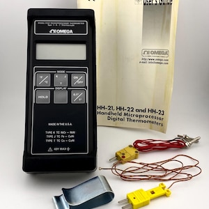 May include: A black handheld digital thermometer with a yellow and red probe. The thermometer has a display showing the temperature in degrees Fahrenheit and Celsius. The device is labeled "OMEGA" and "MODEL HH21 MICROPROCESSOR THERMOMETER".