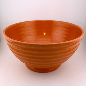 May include: An orange ceramic bowl with a wide, round shape. The bowl has a textured, ridged exterior and a smooth interior. The bowl sits on a small, circular base. The colour is a warm, earthy orange.
