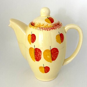 May include: A cream-colored ceramic teapot with a lid, decorated with red and yellow stylized leaf designs.