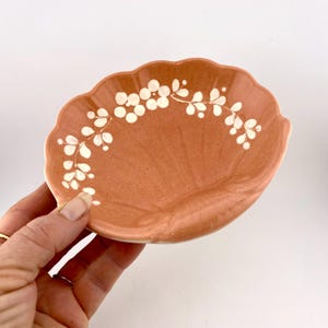 May include: A small, peach-colored ceramic dish shaped like a seashell with a white floral design around the rim.