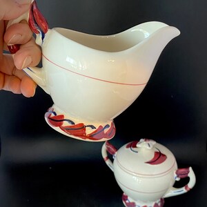 May include: A white ceramic creamer and sugar bowl set with red and blue floral designs. The creamer has a handle shaped like a leaf. The sugar bowl has a lid with a floral design.