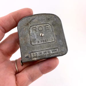 May include: A vintage metal tape measure with the brand name "ROE" and the text "6 FEET". The tape measure also has the text "+2 IN OR 51 MM FOR INSIDE MEASURE". The tape measure is gray and has a metal hook.