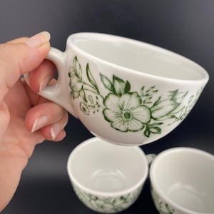Puede incluir: Tazas de té de cerámica blanca con un diseño floral verde. Las tazas tienen un asa y están decoradas con flores y hojas verdes. Las tazas de té están apiladas. Adecuadas para té o café.