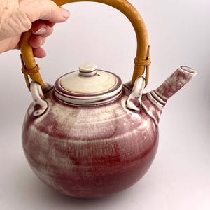 May include: A round, ceramic teapot with a burgundy and cream glaze. It features a light brown, arched handle made of woven material and a matching lid. The spout is striped, and the teapot is held by a hand.