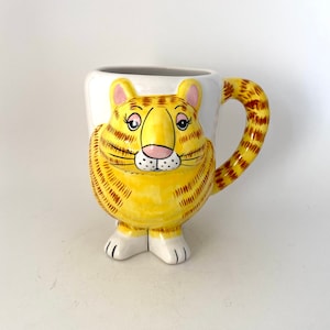 May include: A yellow ceramic mug shaped like a cat with brown stripes. The mug has a white handle and white feet.