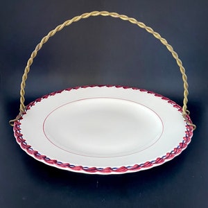 May include: A white ceramic serving plate with a red and blue trim, featuring a gold wire handle.