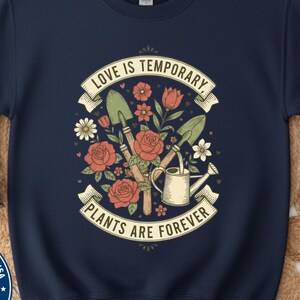 Könnte beinhalten: Marineblaues Sweatshirt mit floralem Design und Gartengeräten. Die Grafik zeigt Rosen, Gänseblümchen, eine Gießkanne und den Text "LOVE IS TEMPORARY, PLANTS ARE FOREVER". Ein kreisförmiges Emblem mit der amerikanischen Flagge befindet sich unten links.