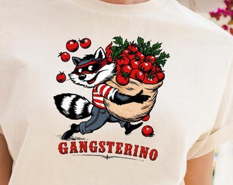 Puede incluir: Una camiseta de color crema con un gráfico de un mapache con una camisa de rayas rojas y blancas y una máscara negra, cargando un saco de tomates rojos. El texto GANGSTERINO está impreso en rojo debajo de la ilustración del mapache.