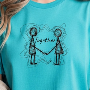 Op de afbeelding: Een turquoise t-shirt met een zwart-witte illustratie van twee handen vasthoudende figuren, met het woord "Together" erboven. Het ontwerp is omgeven door gekrabbelde lijnen. Het shirt is gemaakt van een zacht materiaal.