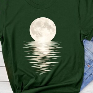 Könnte beinhalten: Ein dunkelwaldgrünes T-Shirt mit kurzen Ärmeln und einem Grafikdruck eines großen, runden, blassweißen Vollmonds in der Mitte der Brust, mit einer stilisierten Spiegelung des Mondes in horizontalen Linien direkt darunter.