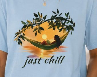 Op de afbeelding: Een lichtblauw t-shirt met korte mouwen en een grafische print van een persoon die rust in een hangmat tussen bladerrijke takken boven een oceaan bij zonsondergang, met de tekst "just chill" in een donker cursief lettertype eronder.