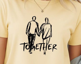 Op de afbeelding: Een lichtgeel T-shirt met korte mouwen en een zwarte lijntekening van twee figuren die elkaars hand vasthouden. Onder de figuren staat het woord TOGETHER in een vet, handgetekend lettertype. Het T-shirt wordt gedragen met een lichtblauw spijkerjasje.