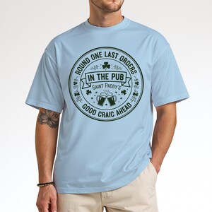 Light Blue unisex t-shirt featuring a vintage green circular Irish pub logo design. The text reads "Round One Last Orders / In The Pub / Saint Paddy's / Good Craic Ahead" surrounding two clinking beer mugs and shamrocks. Clean, classic look for St. Patrick's Day.