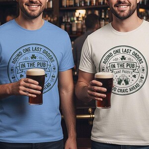 Carolina Blue and Ice Grey Colors unisex t-shirts featuring a vintage green circular Irish pub logo design. The text reads "Round One Last Orders / In The Pub / Saint Paddy's / Good Craic Ahead" surrounding two clinking beer mugs and shamrocks. Clean, classic look for St. Patrick's Day.