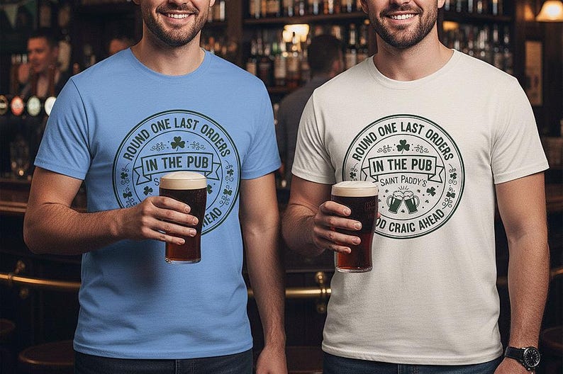 Carolina Blue and Ice Grey Colors unisex t-shirts featuring a vintage green circular Irish pub logo design. The text reads "Round One Last Orders / In The Pub / Saint Paddy's / Good Craic Ahead" surrounding two clinking beer mugs and shamrocks. Clean, classic look for St. Patrick's Day.