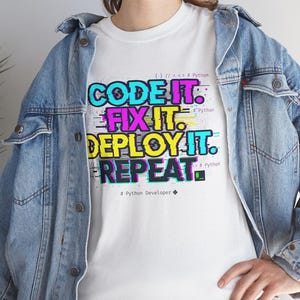 White t-shirt with a neon glitch typography design: "CODE IT. FIX IT. DEPLOY IT. REPEAT." and "# Python Developer". Gritty, modern streetwear for programmers and engineers.