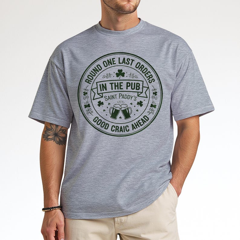 May include: A gray t-shirt with a circular green graphic. The graphic reads "Round One Last Orders In The Pub Saint Paddy's Good Craic Ahead." The design includes shamrocks and beer mugs, perfect for St. Patrick's Day.