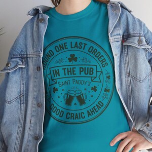 Tropical Blue unisex t-shirt featuring a vintage green circular Irish pub logo design. The text reads "Round One Last Orders / In The Pub / Saint Paddy's / Good Craic Ahead" surrounding two clinking beer mugs and shamrocks. Clean, classic look for St. Patrick's Day.