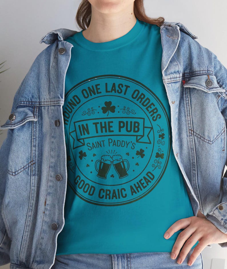 Tropical Blue unisex t-shirt featuring a vintage green circular Irish pub logo design. The text reads "Round One Last Orders / In The Pub / Saint Paddy's / Good Craic Ahead" surrounding two clinking beer mugs and shamrocks. Clean, classic look for St. Patrick's Day.