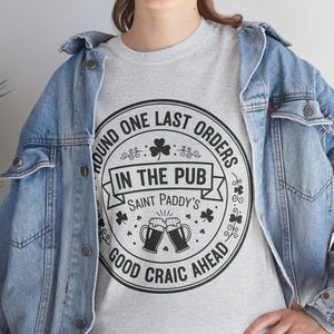 Ash Grey unisex t-shirt featuring a vintage green circular Irish pub logo design. The text reads "Round One Last Orders / In The Pub / Saint Paddy's / Good Craic Ahead" surrounding two clinking beer mugs and shamrocks. Clean, classic look for St. Patrick's Day.