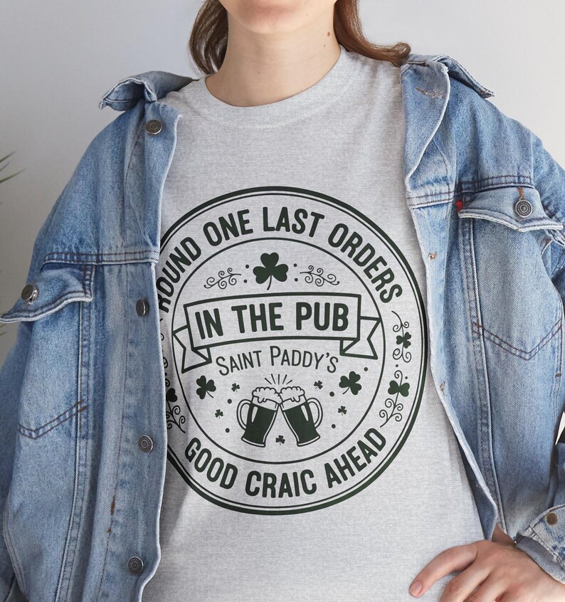 Ash Grey unisex t-shirt featuring a vintage green circular Irish pub logo design. The text reads "Round One Last Orders / In The Pub / Saint Paddy's / Good Craic Ahead" surrounding two clinking beer mugs and shamrocks. Clean, classic look for St. Patrick's Day.