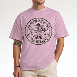 Light pink unisex t-shirt featuring the "Good Craic Ahead" pub seal design in dark green. A fun, playful twist on traditional St. Patrick's Day gear, perfect for women or men who want a softer color palette while keeping the Irish drinking humor.
